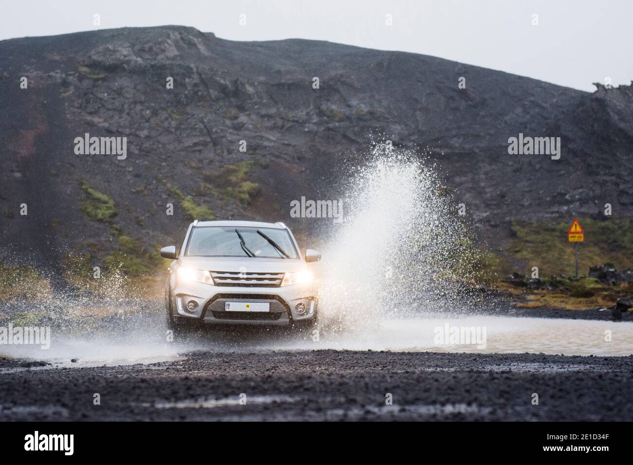A silver coloured SUV races across a pond on a four wheel drive black road, spraying water up to twice its height, in South Iceland. Stock Photo