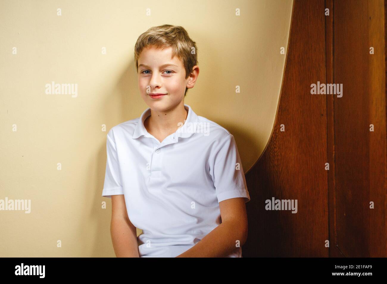 A young boy sits on wooden bench leaning against wall with direct gaze Stock Photo