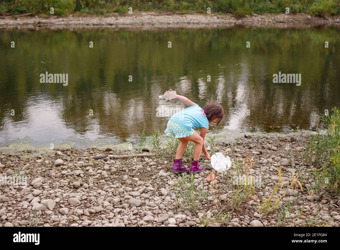 A little girl cleans litter up off of the shoreline of a river Stock Photo