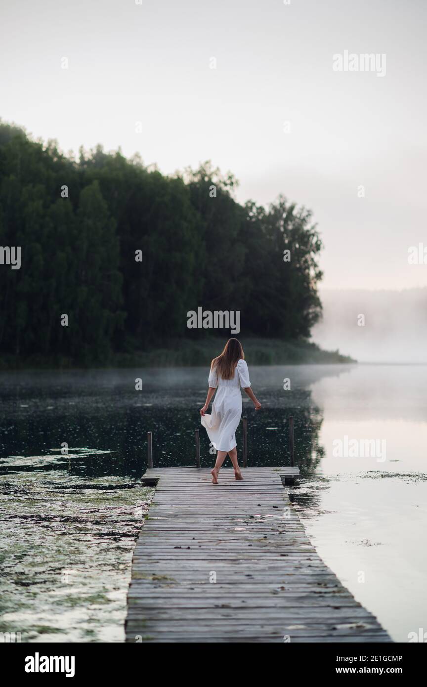 Young woman in white dress on a lake at chilly morning with a mist over water. Stock Photo