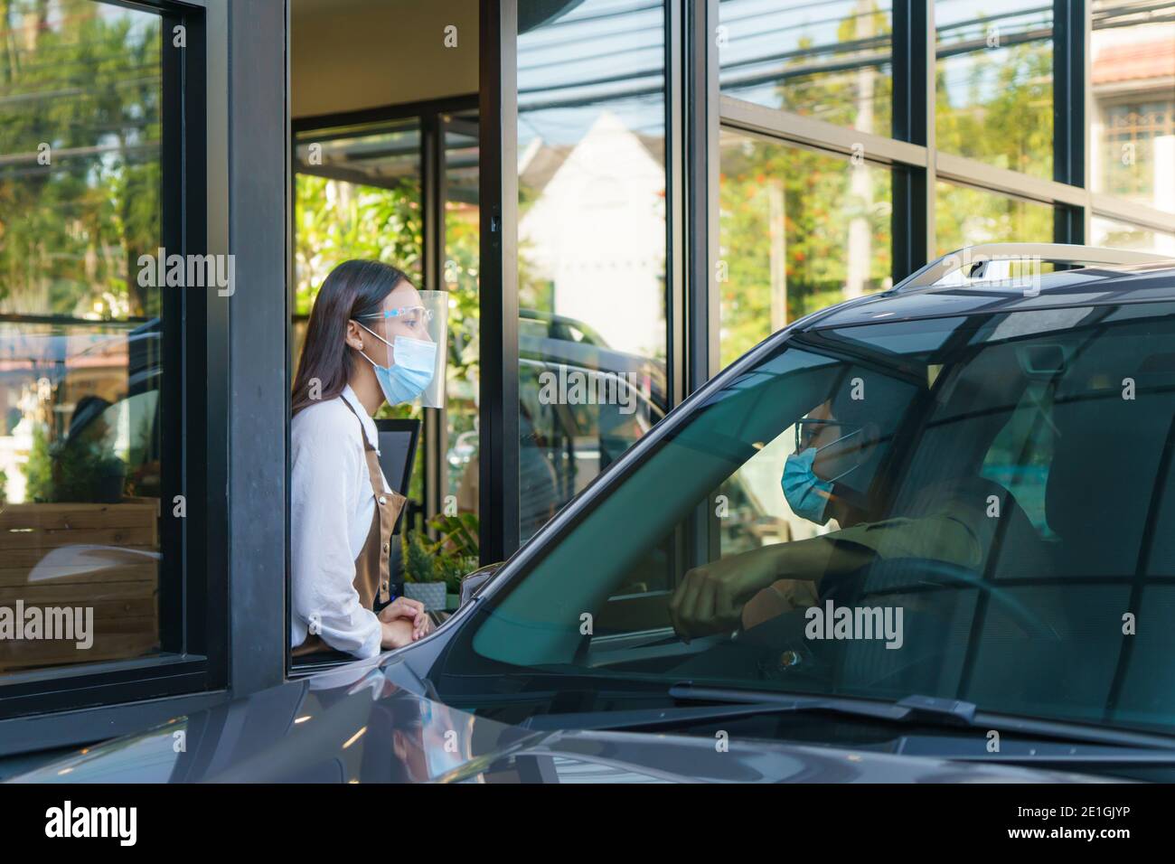 Asian man in protective mask order food and coffee with woman waitress wearing face mask and face shield at drive thru during coronavirus outbreak. Stock Photo