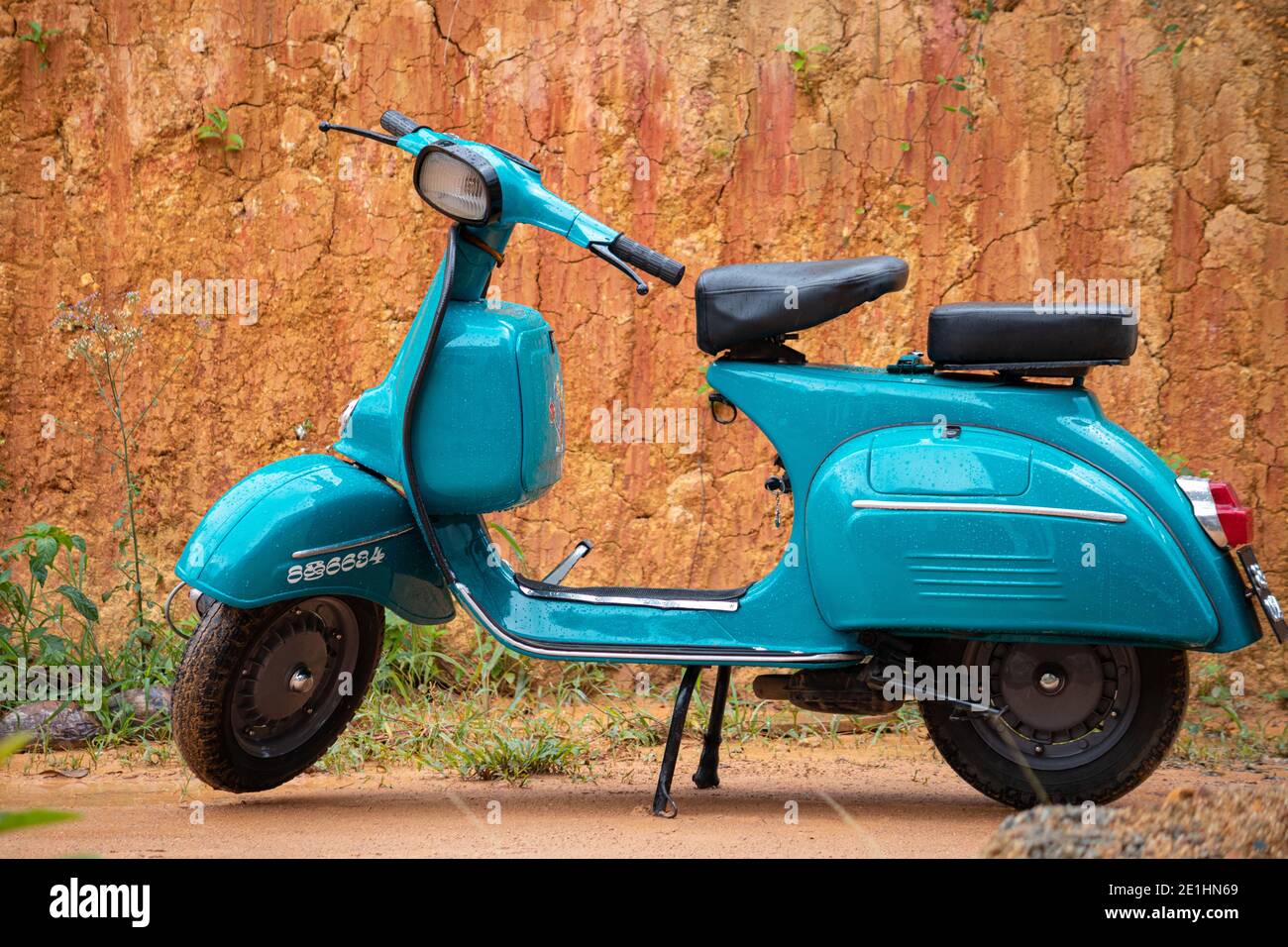 Old vintage scooter motorbike in display against clay wall. light blue color and two-seaters, compartments for storage, round headlamp in front. Stock Photo