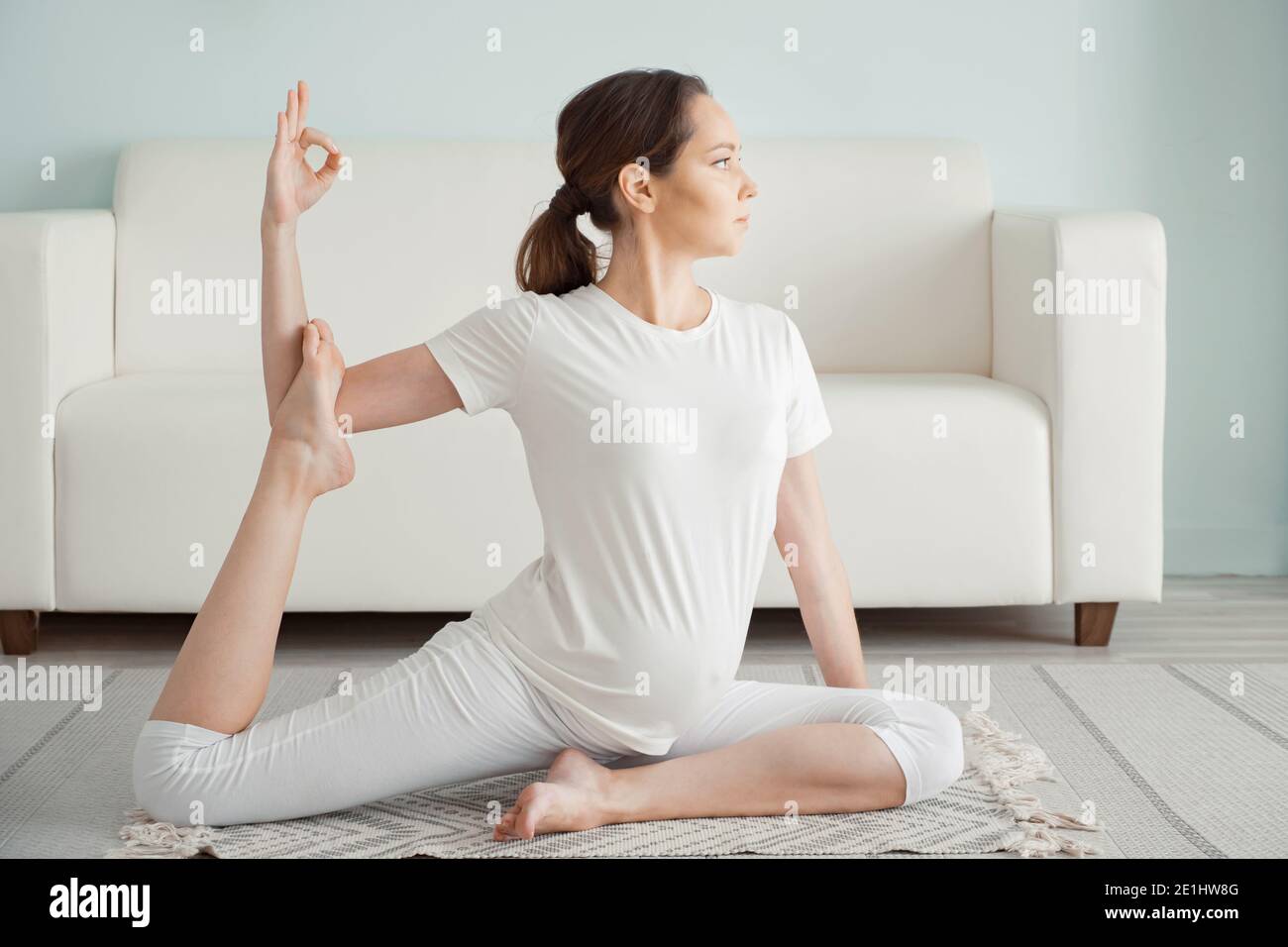 Calm young pregnant brunette woman does ardha matsyendrasana practicing yoga position on floor near sofa at home close-up Stock Photo