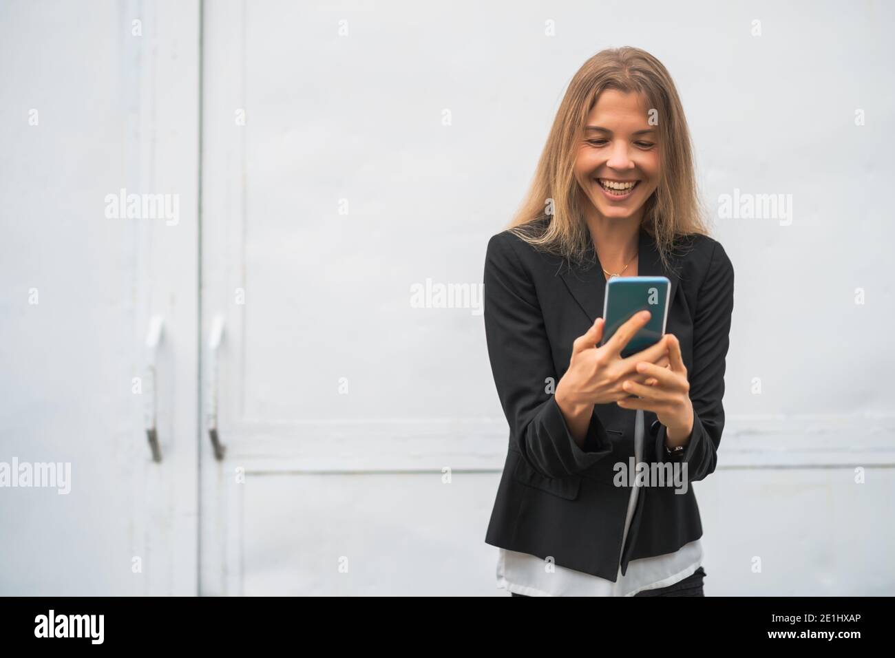 Outdoor portrait of happy modern businesswoman. Blonde businesswoman is standing outdoor and looking at smartphone. Stock Photo