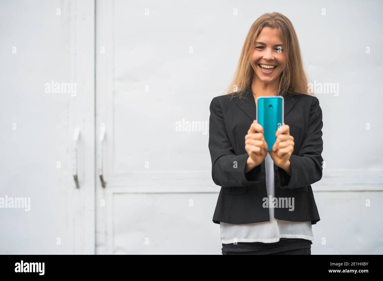 Outdoor portrait of happy modern businesswoman. Blonde businesswoman is standing outdoor and looking at smartphone. Stock Photo