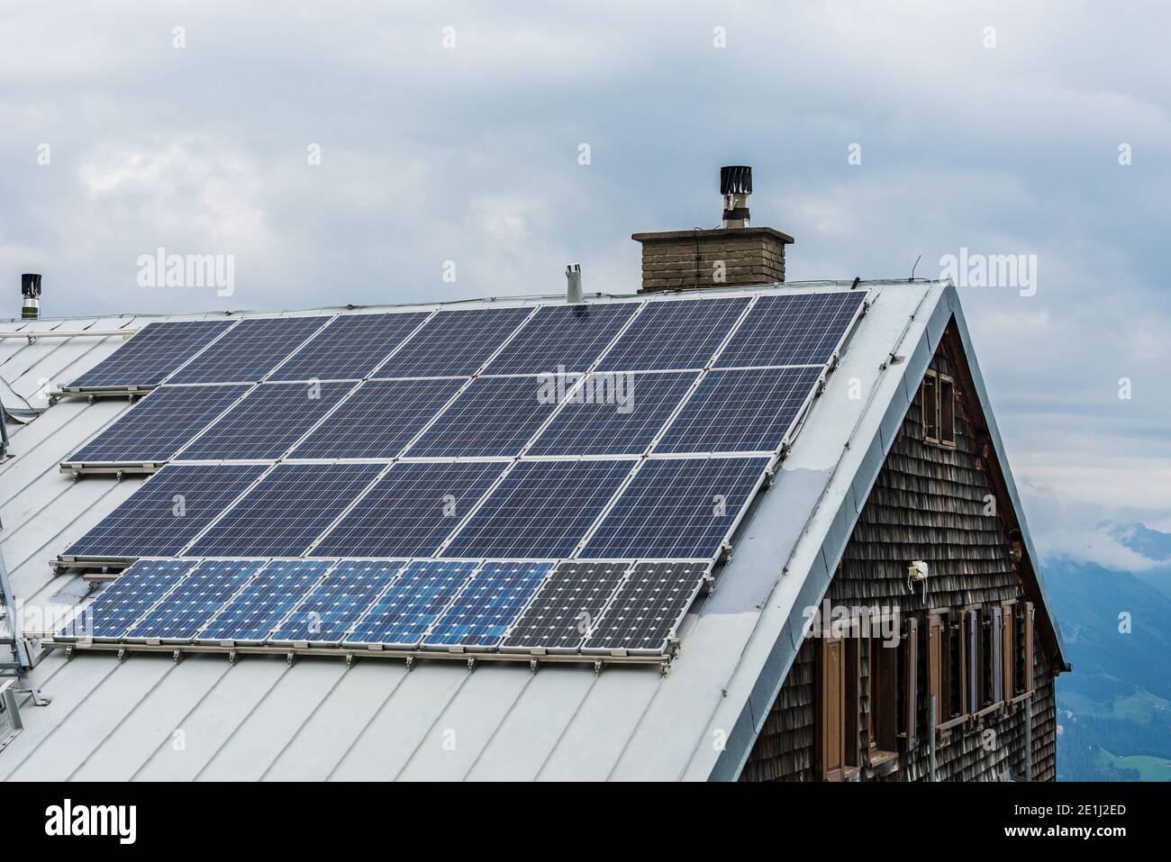 Solar photovoltaic panels PV on a house roof. Electricity from the sun during winter. House with windows and chimney, mountains at the background. Stock Photo