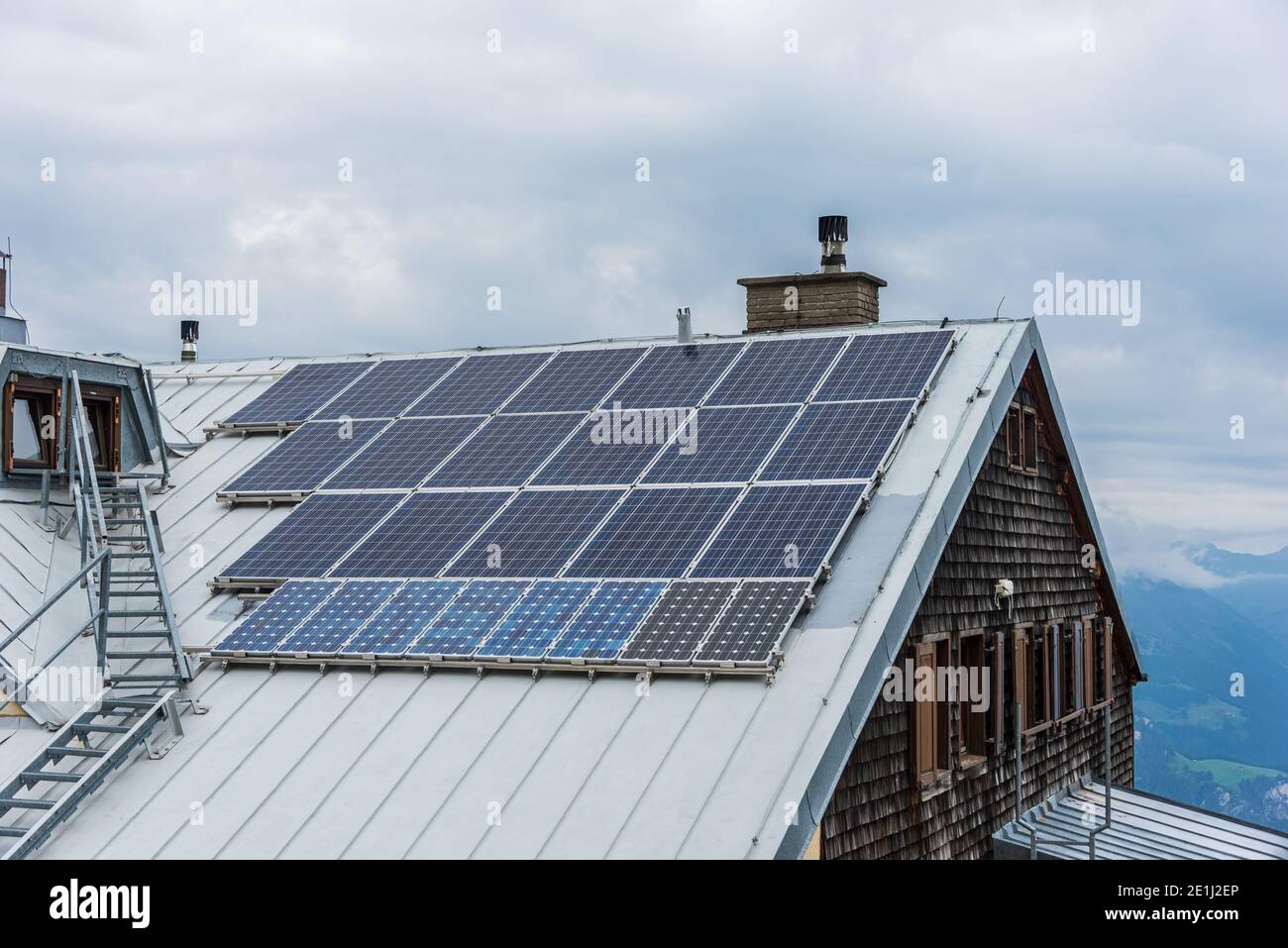 Solar photovoltaic panels PV on a house roof. Electricity from the sun during winter. House with windows and chimney, mountains at the background. Stock Photo
