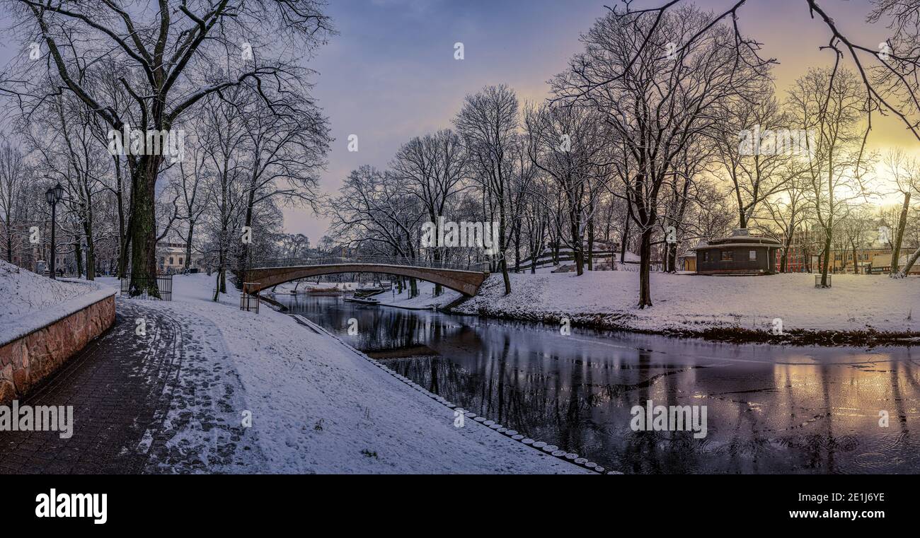 Panoramic winter landscape with sunrise in snowy park with beautiful bridge over small canal, street light and covered in snow tress. View of Bastion Stock Photo