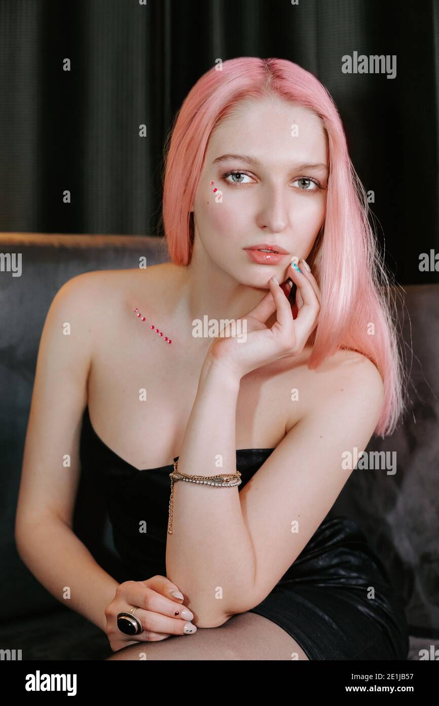 Portrait of a pretty blonde girl wearing a stylish dark strapless dress,  sitting on a couch; she has little makeup jewels as decoration in her face Stock Photo