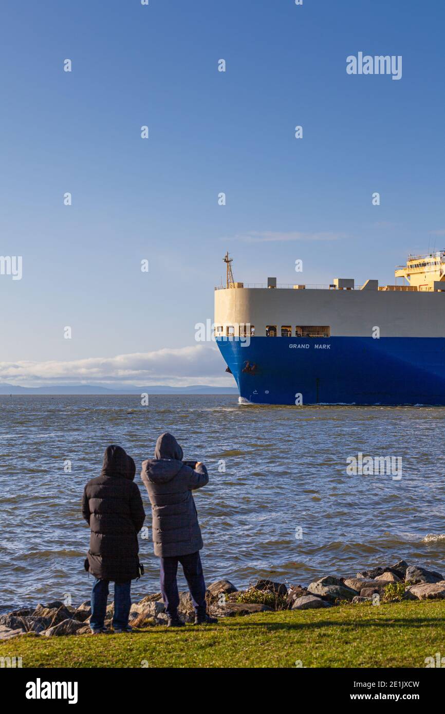 Two young women photograghing the arrival of a large vehicle transport ship in the Fraser River estuary en route from Asia, Steveston British Columbia Stock Photo