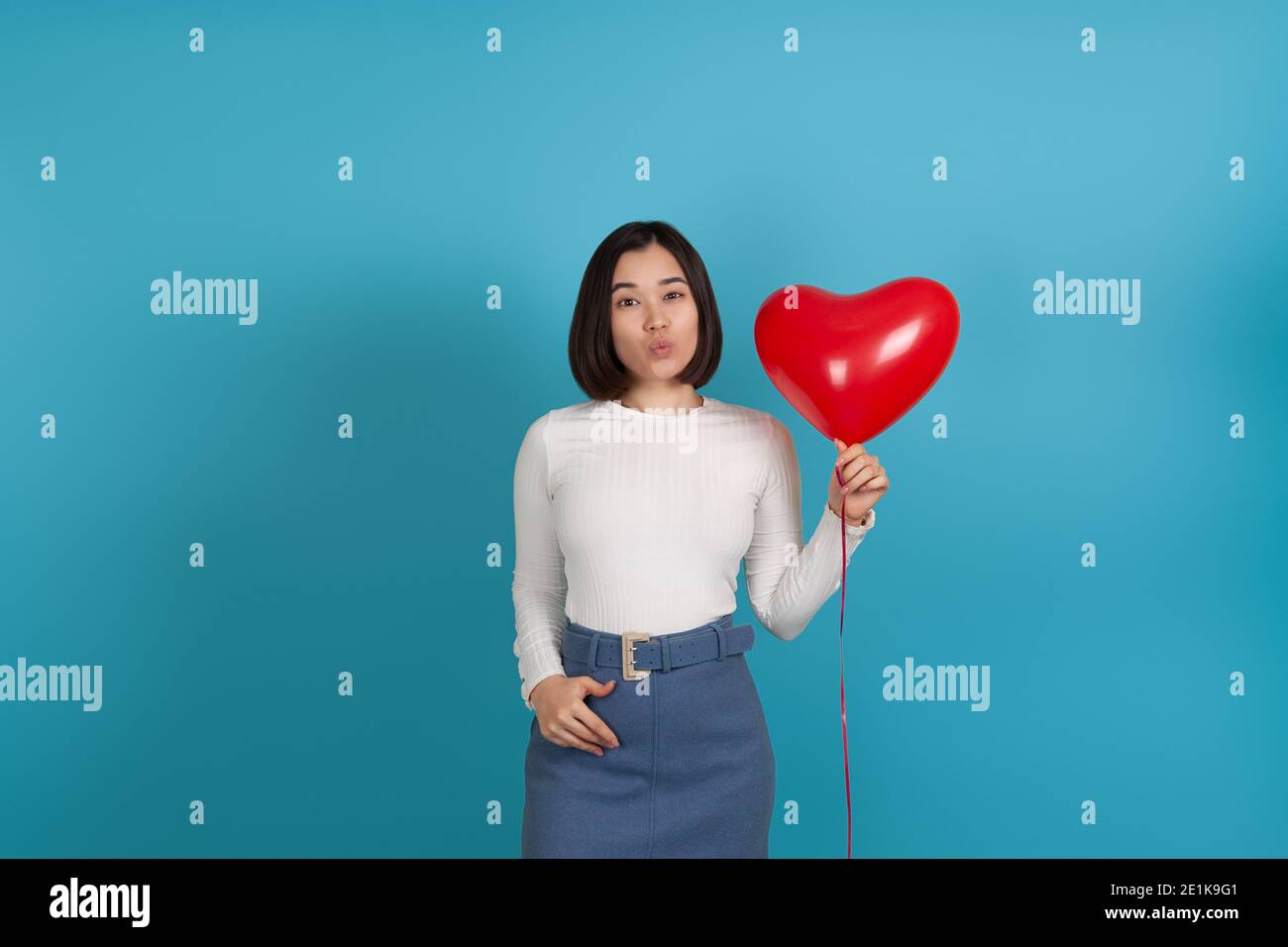 portrait of a young Asian woman blowing a kiss and holding a red heart-shaped balloon isolated on a blue background Stock Photo