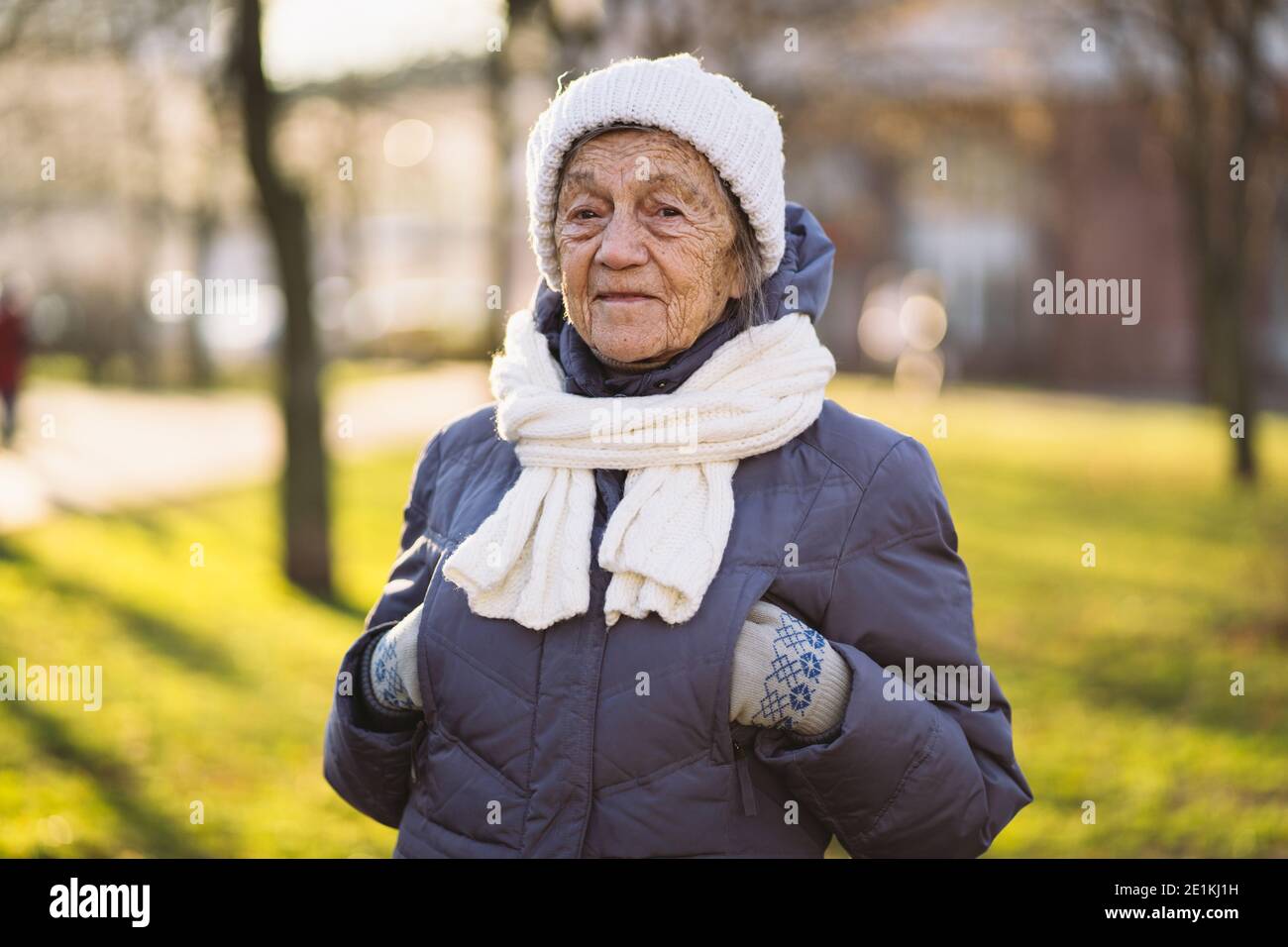 Portrait Caucasian senior woman with gray hair and deep wrinkles 90 years old posing in warm clothes, white knitted scarf and hat in park, sunny frost Stock Photo