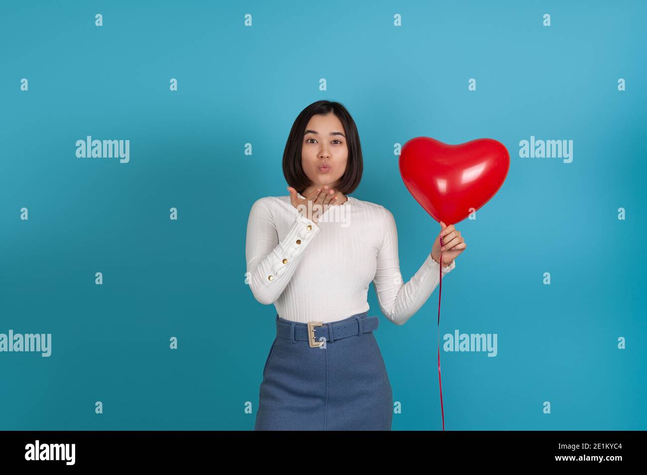 Studio portrait of a young Asian woman blowing a kiss with her palm and holding a red heart-shaped balloon isolated on a blue background Stock Photo