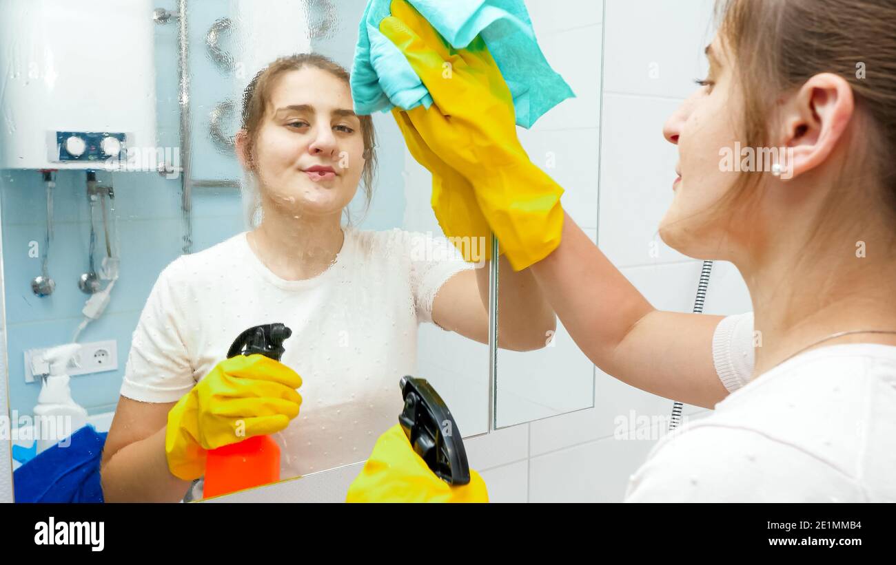 Smiling young woman cleaning and washing bathroom mirror with chemical detergent spray. Housewife doing home cleanup and housekeeping chores Stock Photo