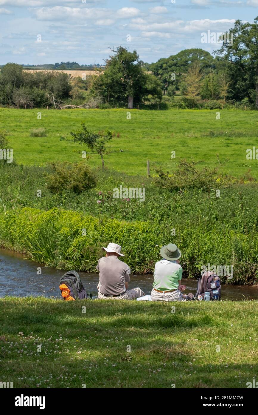 Elderly hiking couple taking a picnic break on the river bank and enjoying nature. Rewe village, Devon, England. Stock Photo
