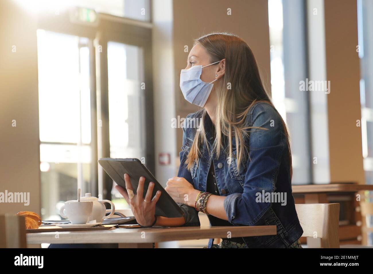 Young woman sitting at coffee shop and working on digital tablet with face mask Stock Photo