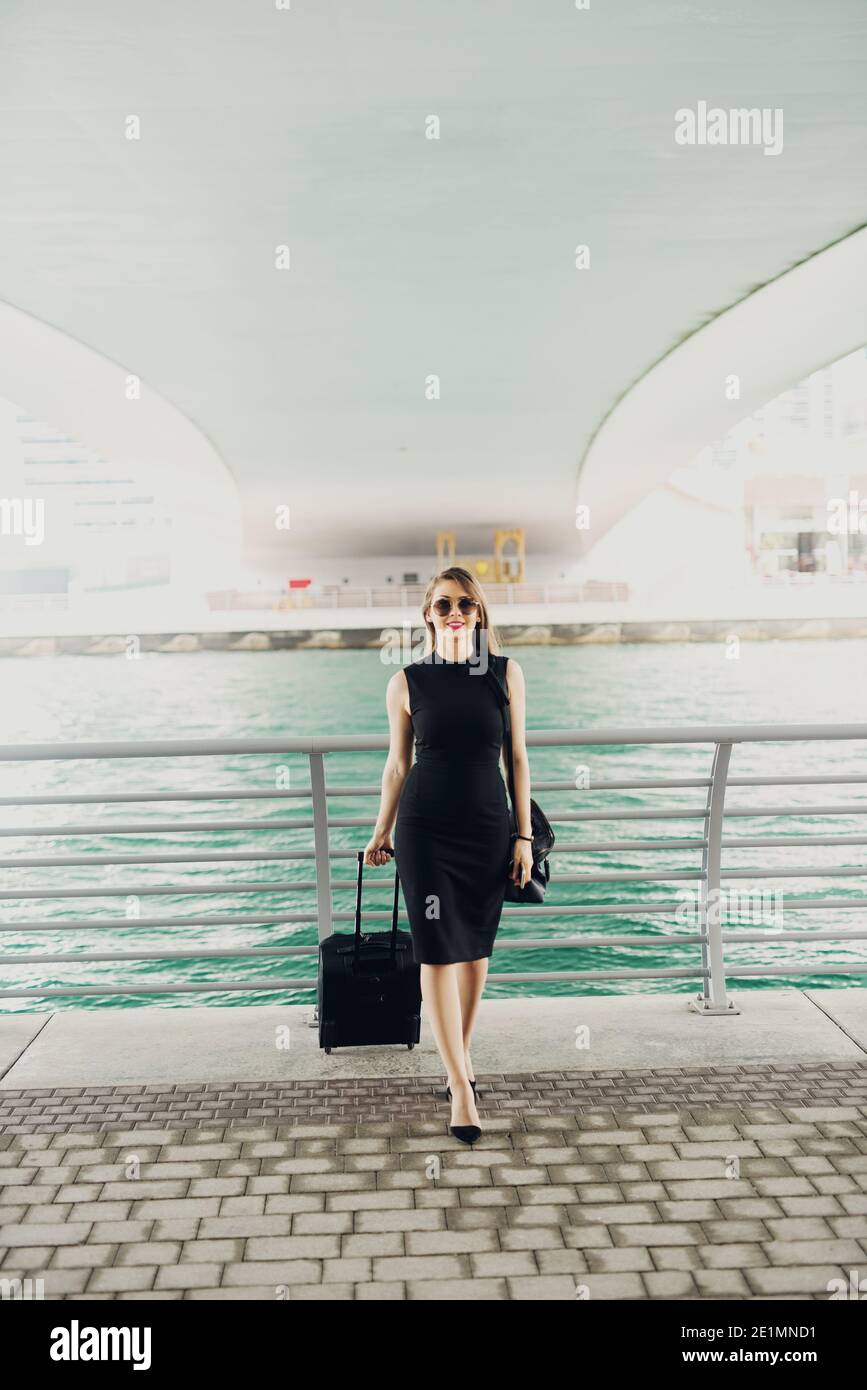 Young modern confident business woman pulling a suitcase in a Dubai Marine. Starting a new job in a big city. Stock Photo