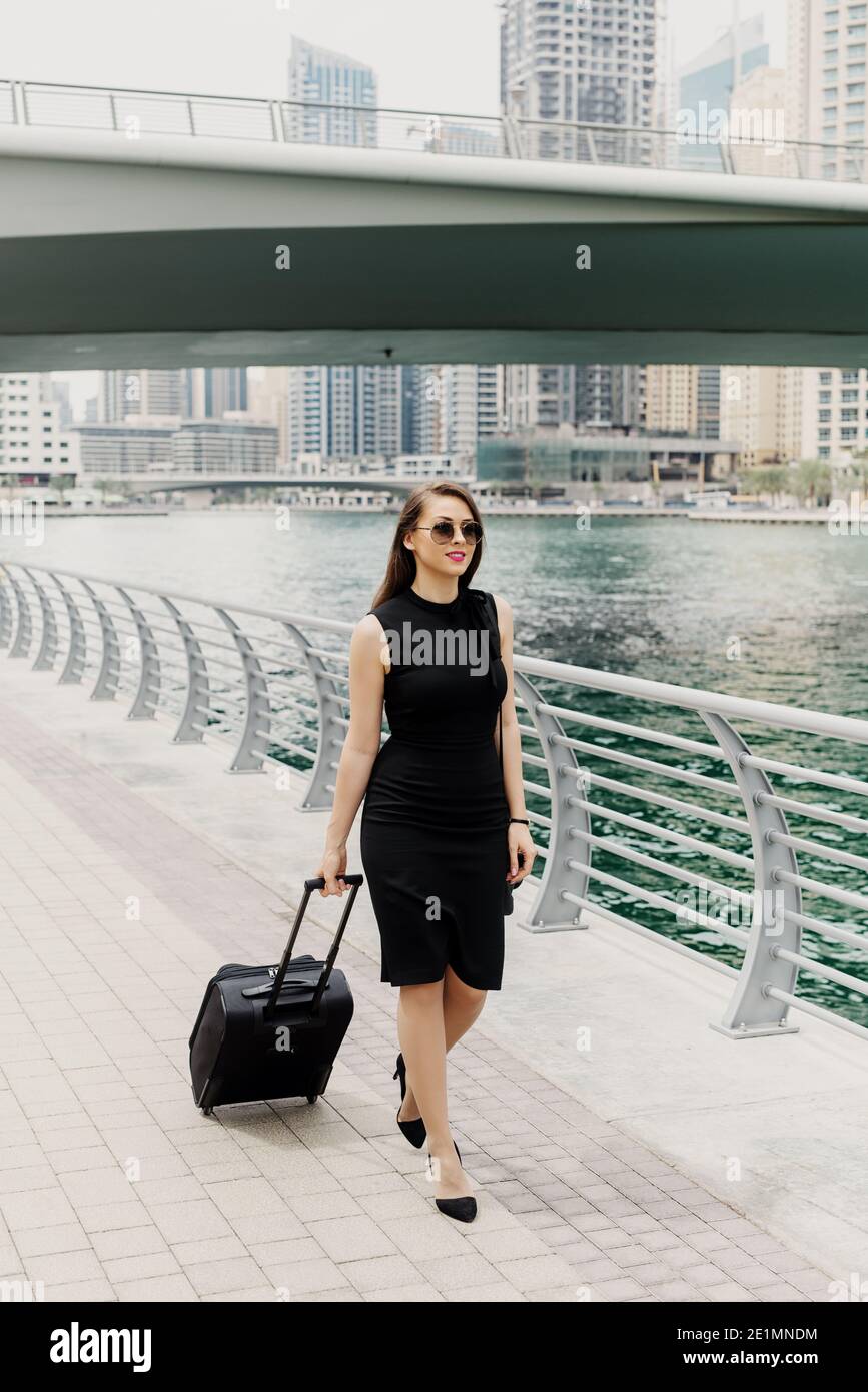 Young modern confident business woman pulling a suitcase in a Dubai Marine. Starting a new job in a big city. Stock Photo