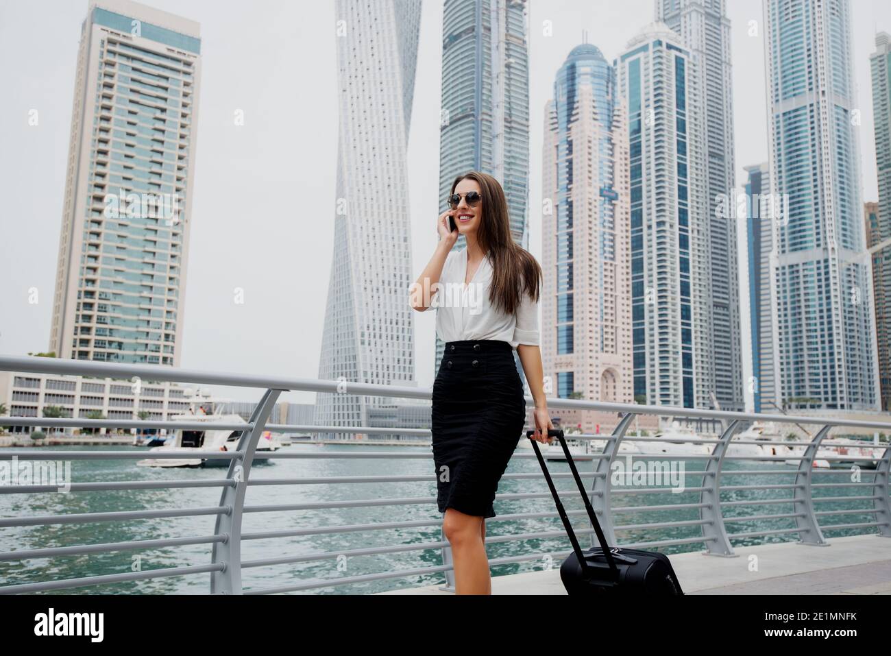 Busy young modern business woman in suit pulling a suitcase in a Dubai Marine. Walking to her office and talking on a telephone.. Stock Photo