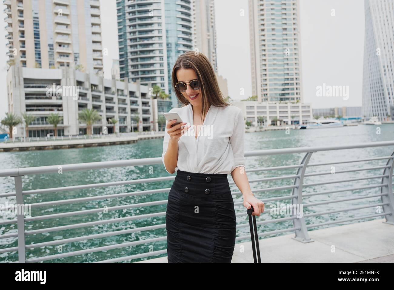 Beautiful young modern businesswoman standing in a Dubai Marine and looking at her telephone with suitcase bar in one hand. Stock Photo