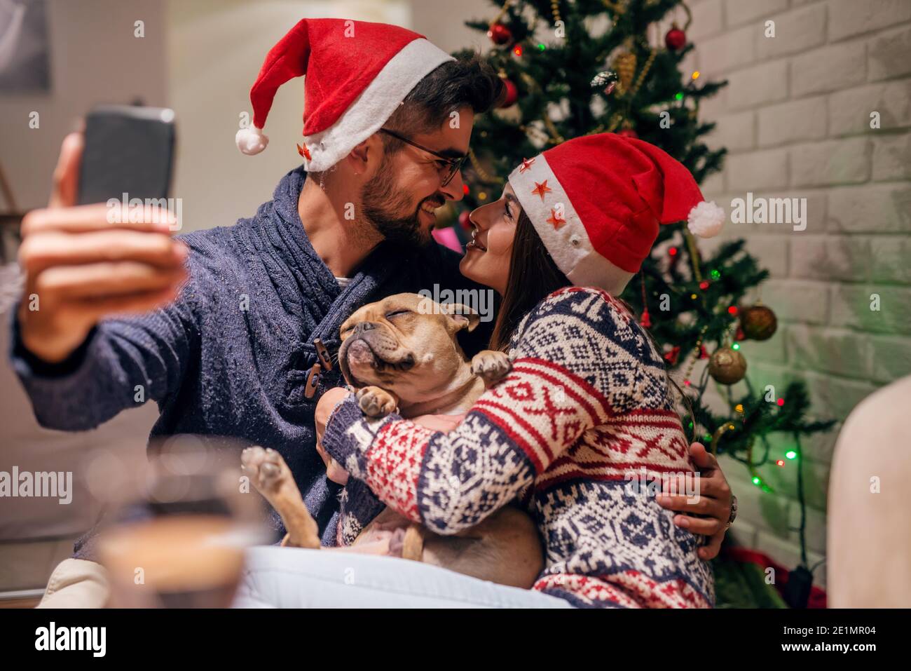 Couple taking self portrait with their dog in front of Christmas tree. On heads santa's hats. Christmas holidays concept. Stock Photo