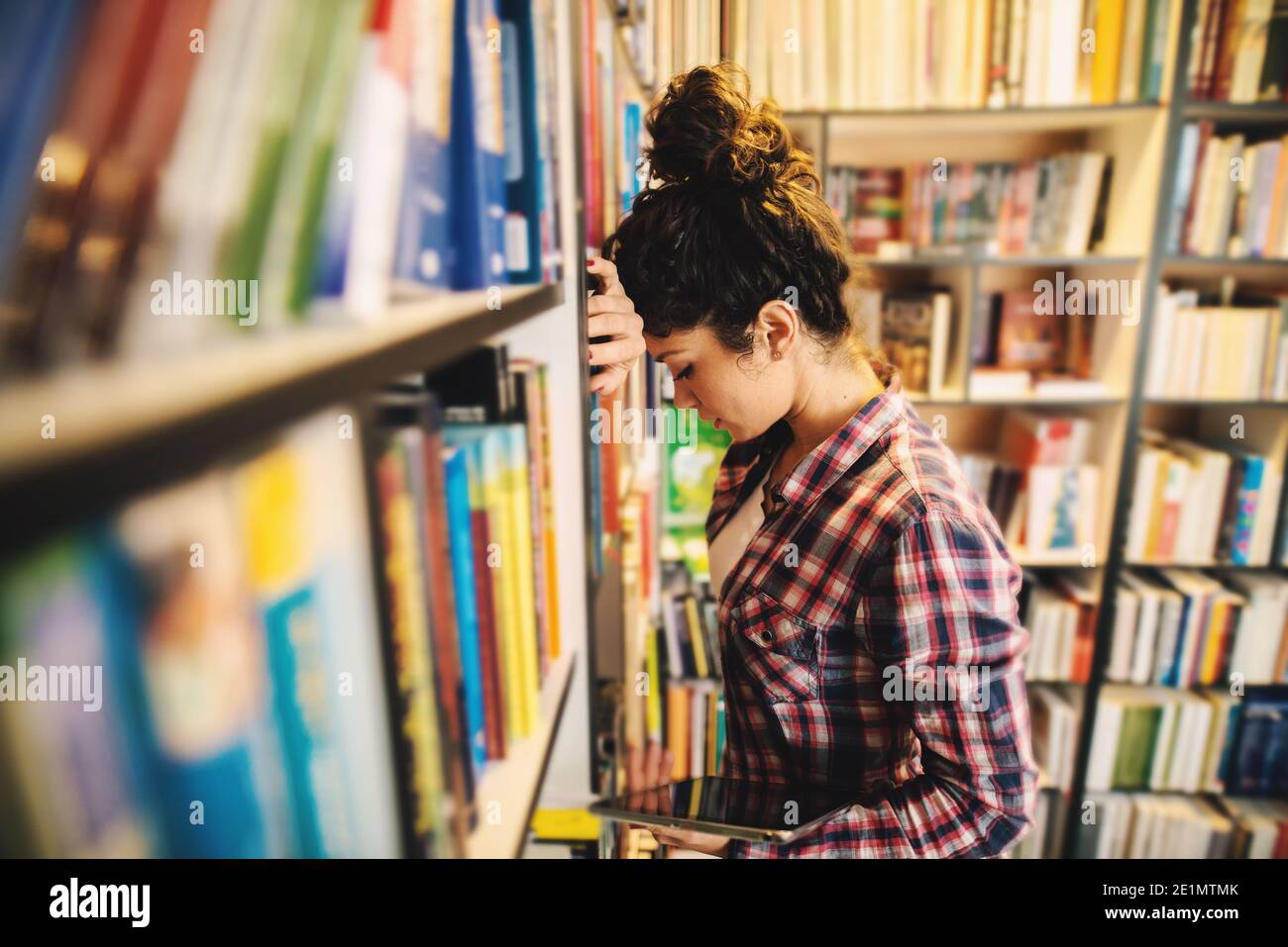 Tired young student girl in library holding tablet in hand and taking few moments to rest from studying. Stock Photo