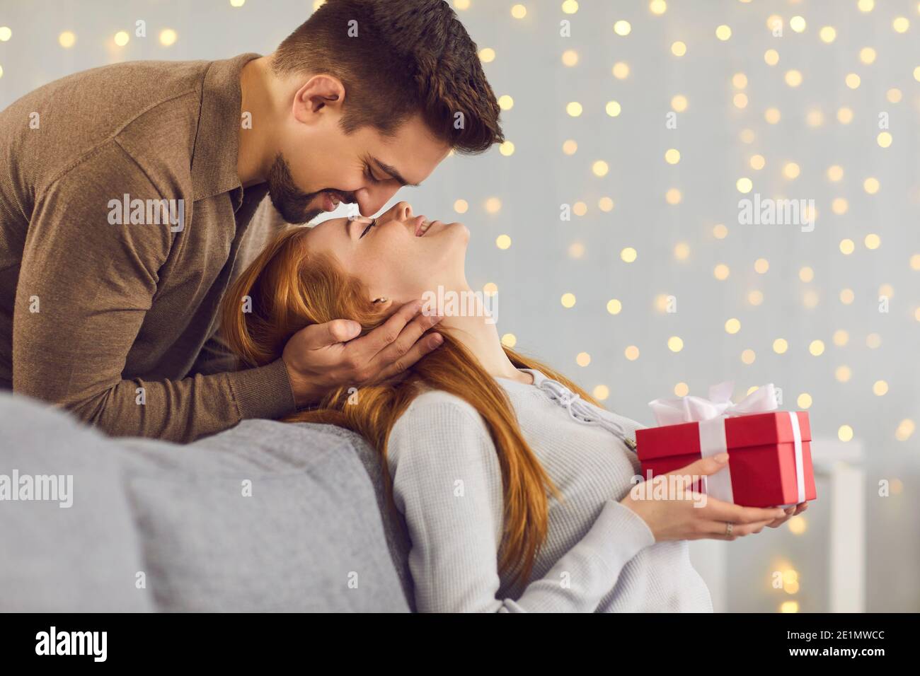 Loving man standing behind and kissing his girlfriend on the forehead, who is sitting on the couch. Stock Photo
