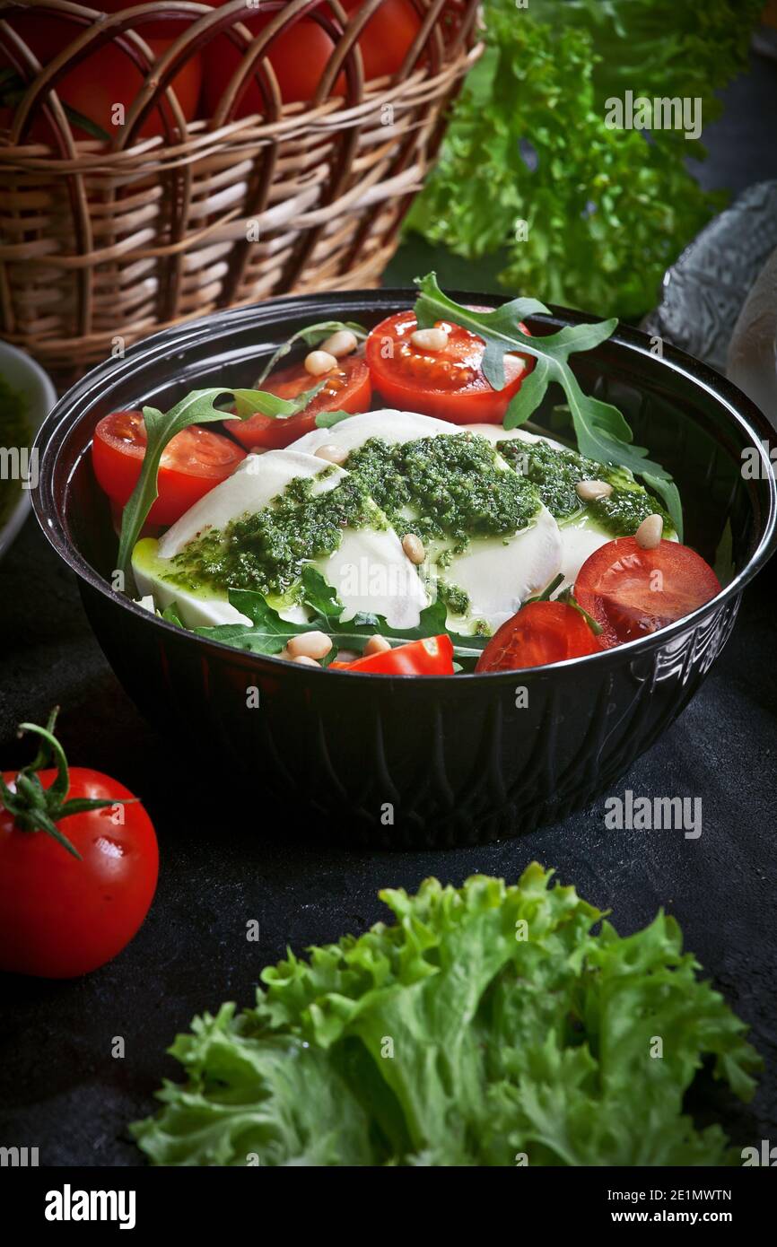 fresh mozzarella salad with lettuce and arugula leaves, pesto sauce, fresh cherry tomatoes and cedar nuts, on a black plate on dark slate table backgr Stock Photo