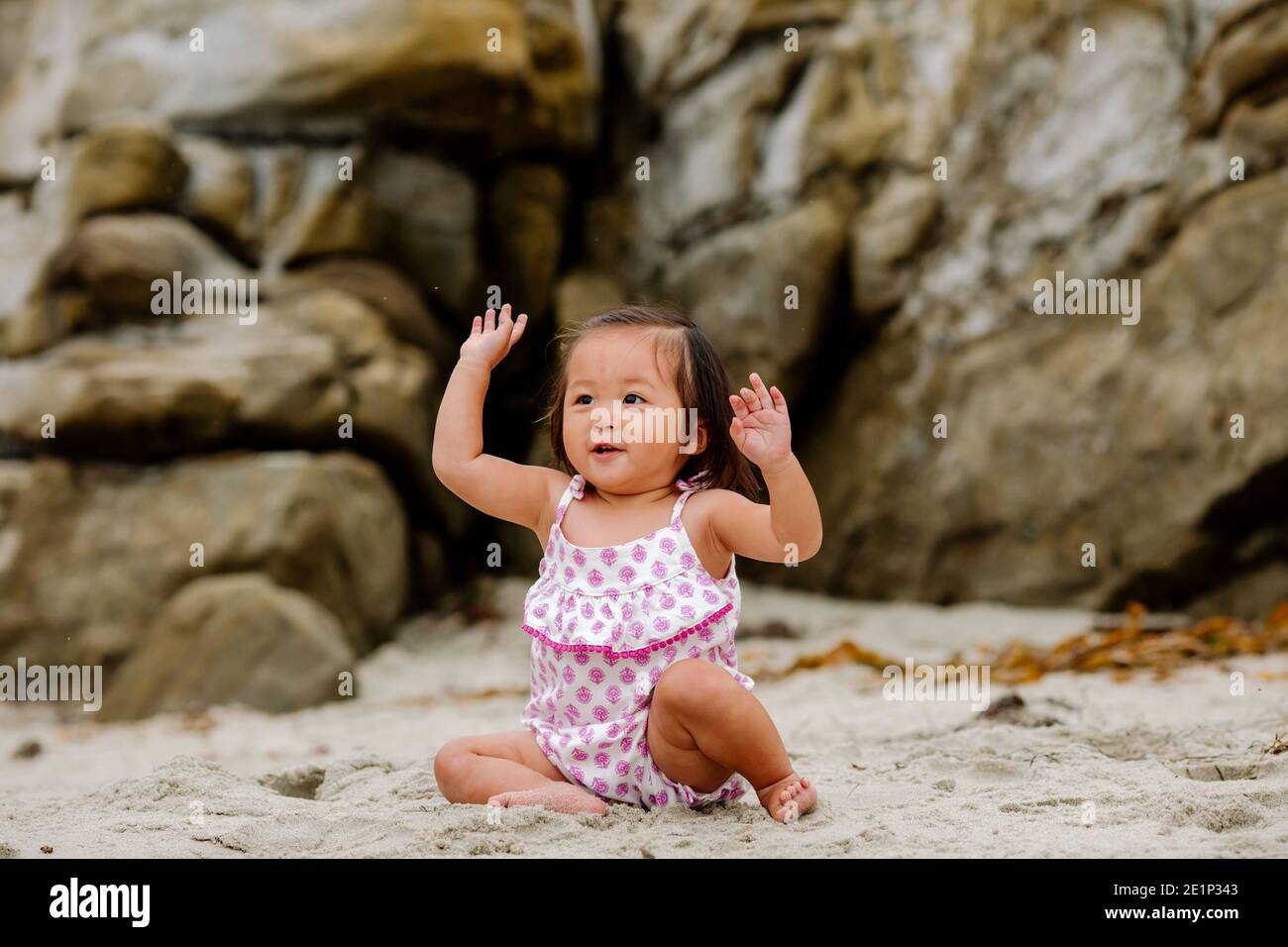 Happy young Asian child with raised arms sitting in sand near rocks Stock Photo