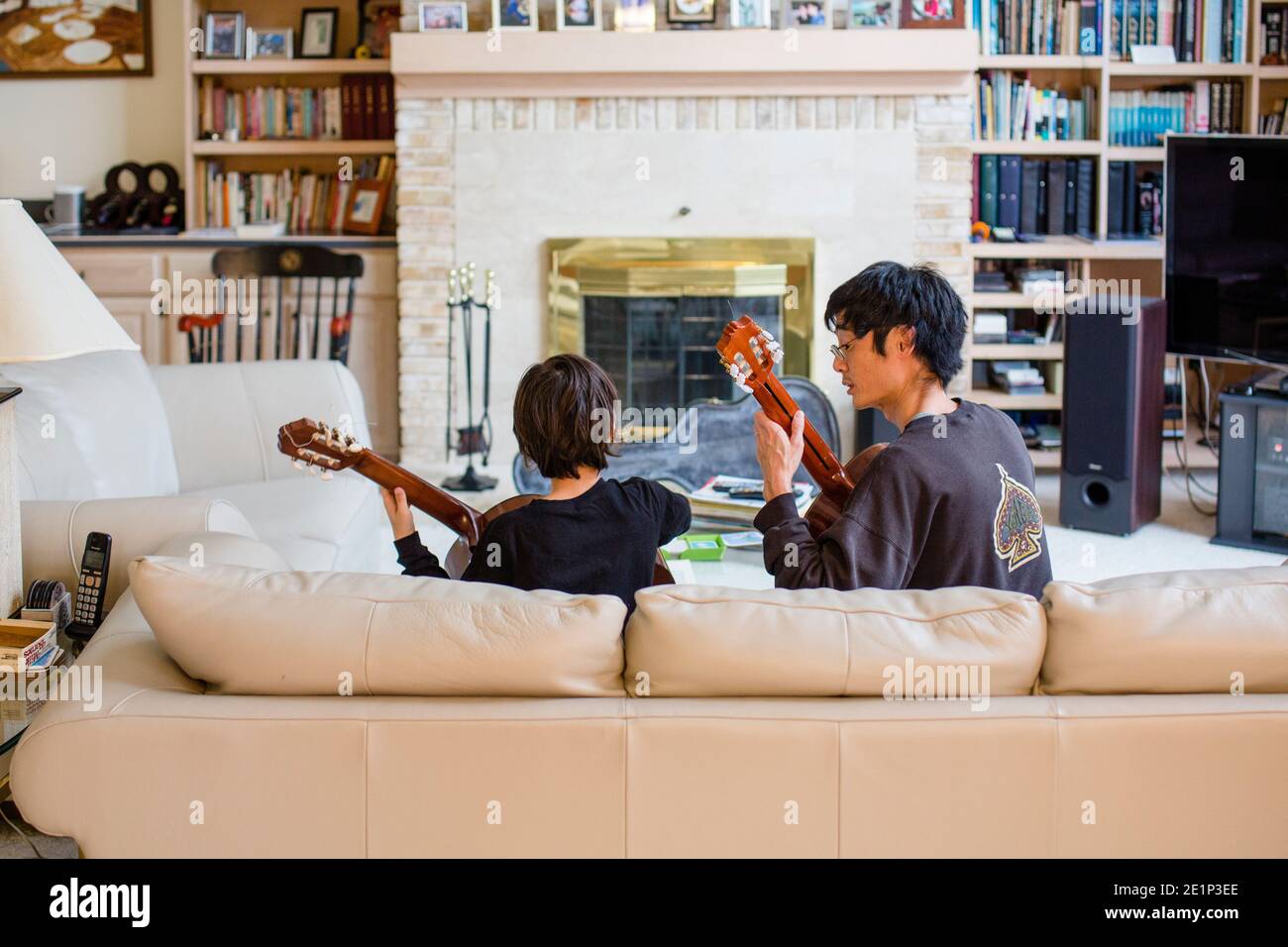 A father and son sit together on couch playing classical guitar music Stock Photo