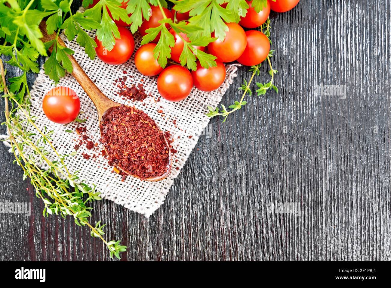 Dried tomato flakes in a spoon on burlap, fresh small tomatoes, parsley and thyme on dark wooden board background from above Stock Photo