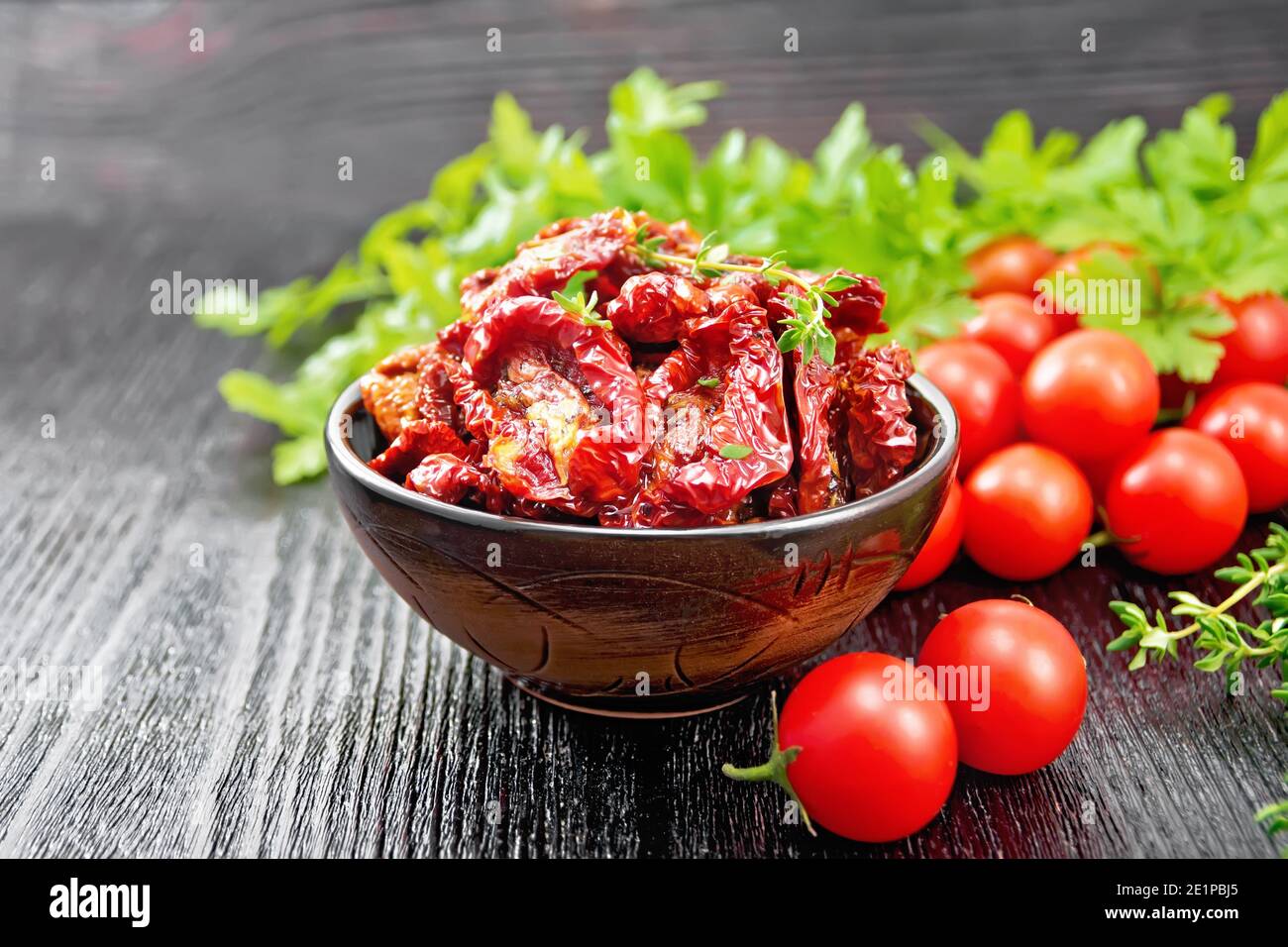 Sun-dried tomatoes in oil with thyme and basil in a bowl, fresh small tomatoes, parsley on dark wooden board background Stock Photo
