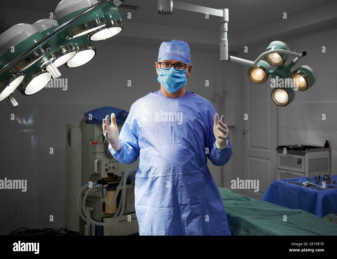 Portrait of surgeon looking at camera while standing in operating room at hospital. Man doctor wearing surgical uniform, sterile gloves, protective face mask, medical cap. Concept of medicine, surgery Stock Photo