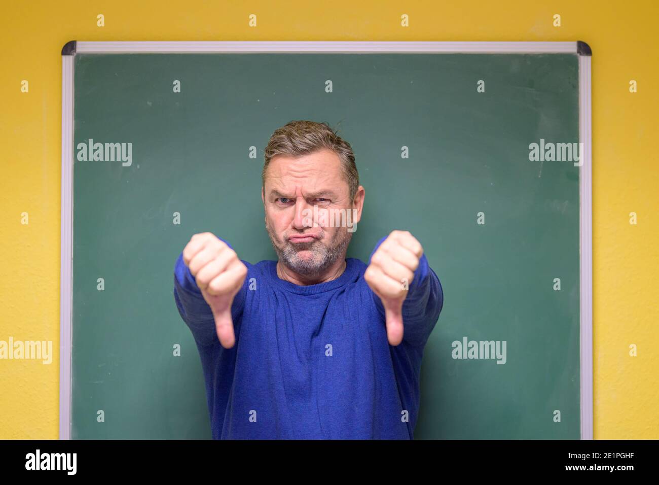 Disappointed male teacher giving a double thumbs down with a grimace as ...
