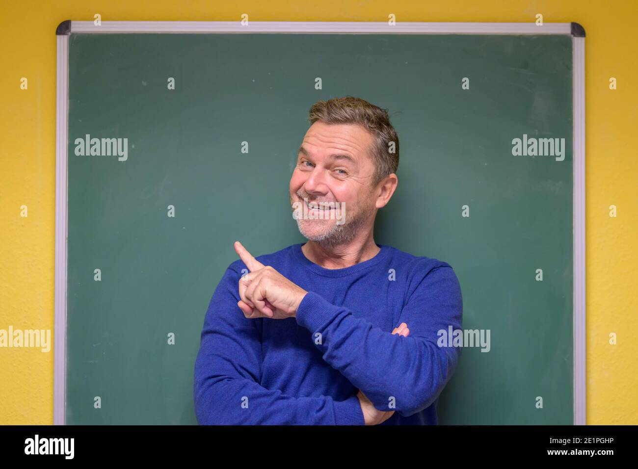 Male teacher pointing to the chalkboard behind him with a beaming smile and look of anticipation as he face s the camera Stock Photo