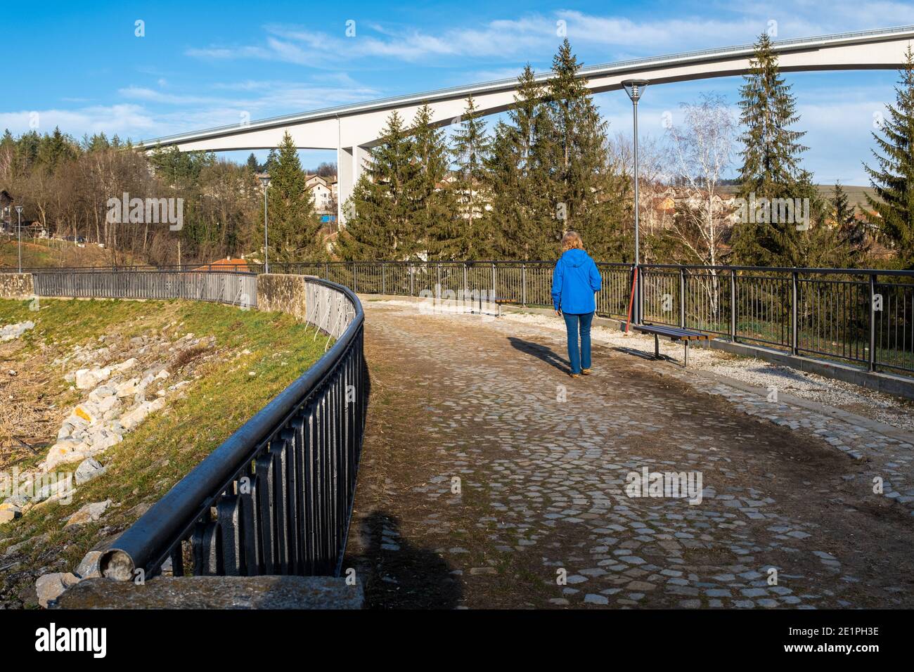 Woman in blue coat walking away on railed dam causeway with concrete cantilever viaduct as background on a sunny winter day. Stock Photo