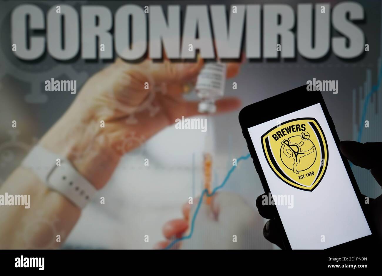 The Burton Albion Football Club logo seen displayed on a mobile phone with a Coronavirus illustration on a monitor in the background Stock Photo