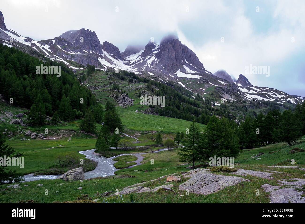 Mountain landscape with a river flowing at the bottom of a valley. In the background, a few clouds veil the mountain tops. Stock Photo