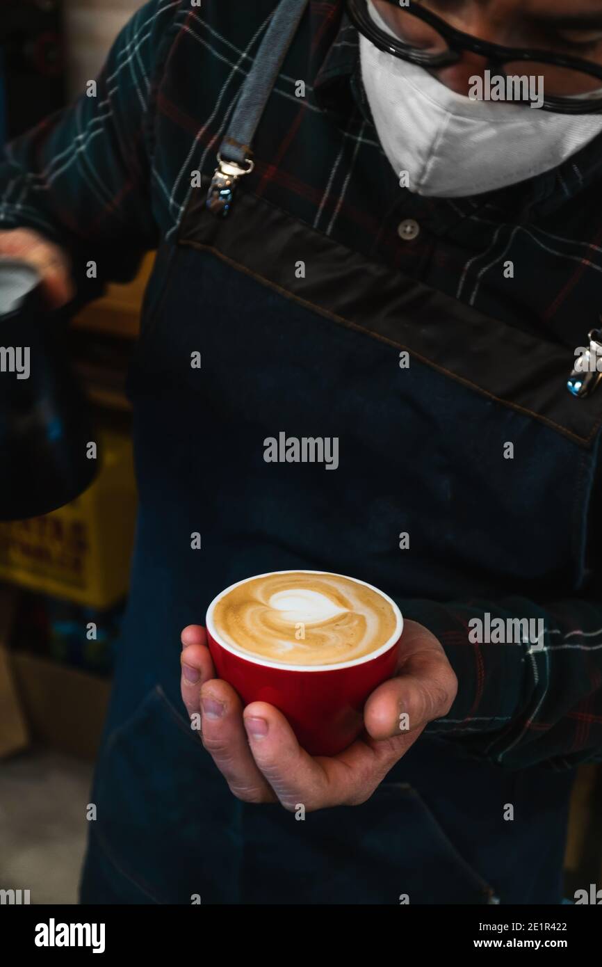 Close-up of a masked unrecognizable barista making a cappuccino in a red cup Stock Photo