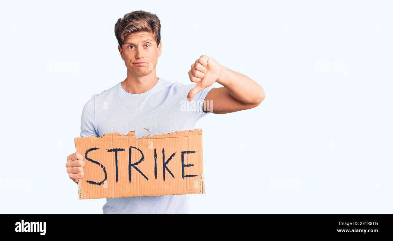 Young handsome man holding strike banner cardboard with angry face, negative sign showing dislike with thumbs down, rejection concept Stock Photo