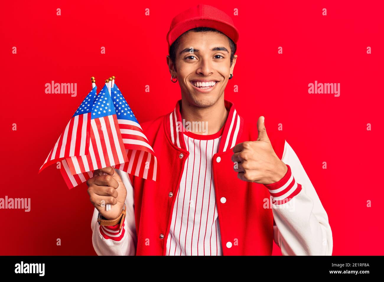 Young african amercian man wearing baseball uniform holding america flags smiling happy and positive, thumb up doing excellent and approval sign Stock Photo