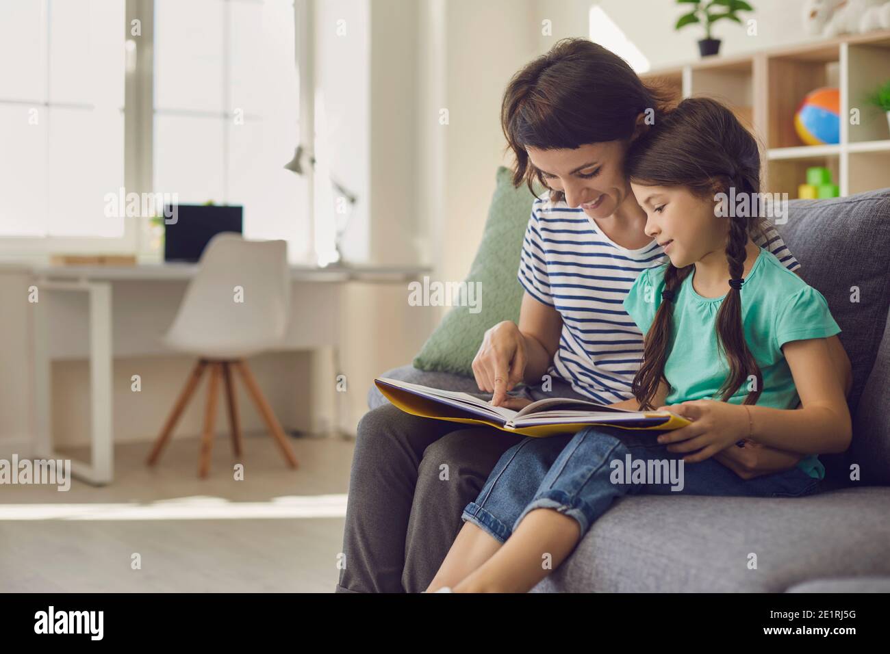 Charming mother is showing images in a book to her cute little daughter at home. Stock Photo