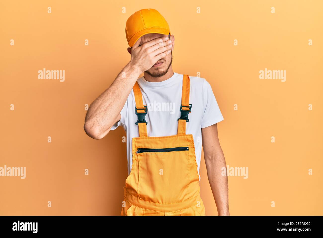 Hispanic young man wearing handyman uniform covering eyes with hand ...