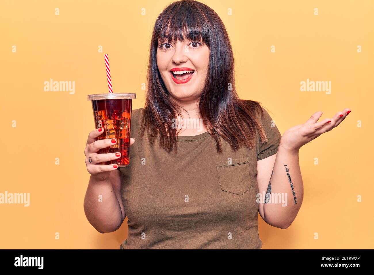 Young plus size woman drinking glass of cola beverage celebrating achievement with happy smile and winner expression with raised hand Stock Photo