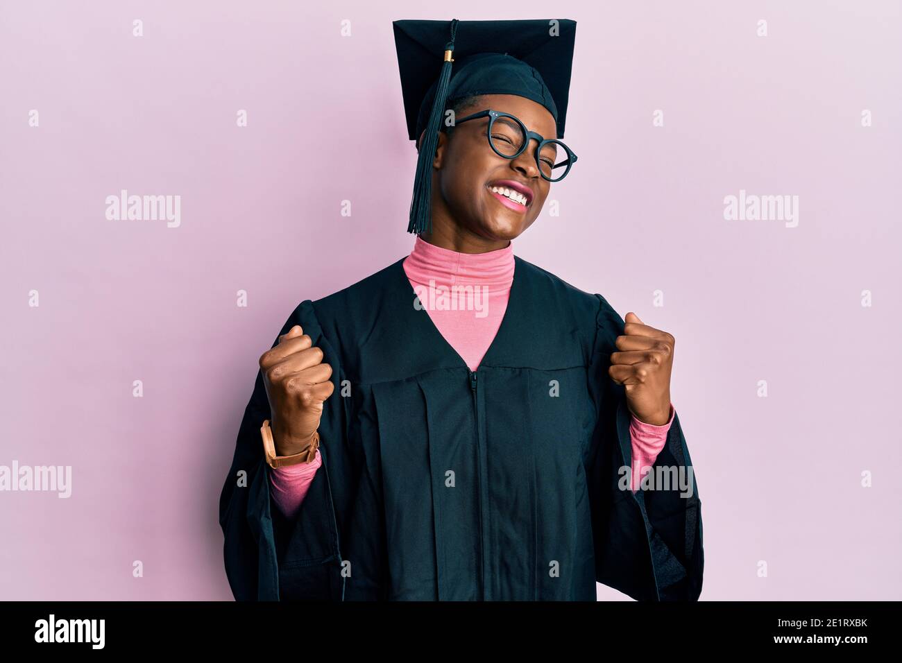 Young african american girl wearing graduation cap and ceremony robe very happy and excited doing winner gesture with arms raised, smiling and screami Stock Photo
