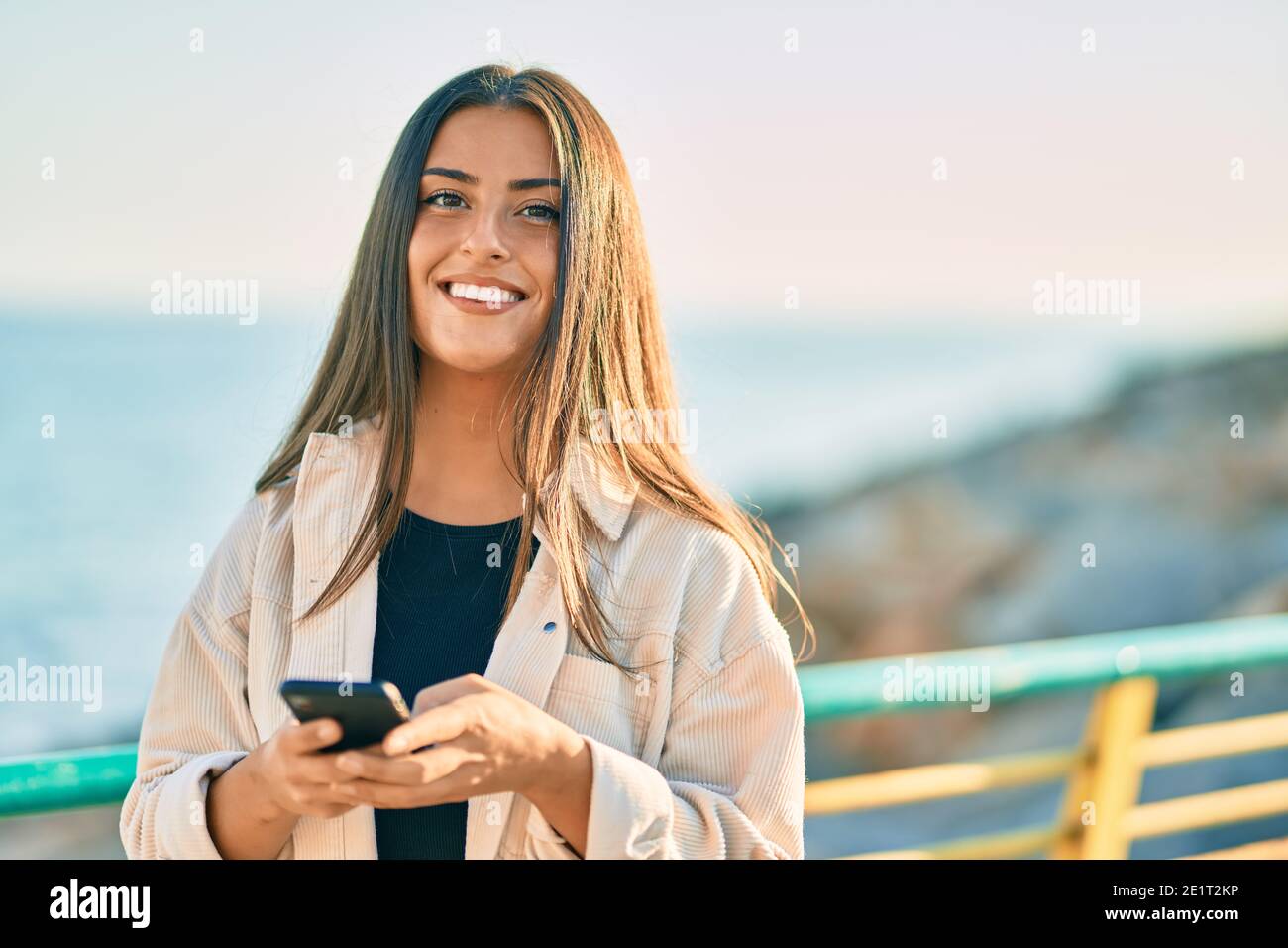 Young hispanic girl smiling happy using smartphone at the promenade ...