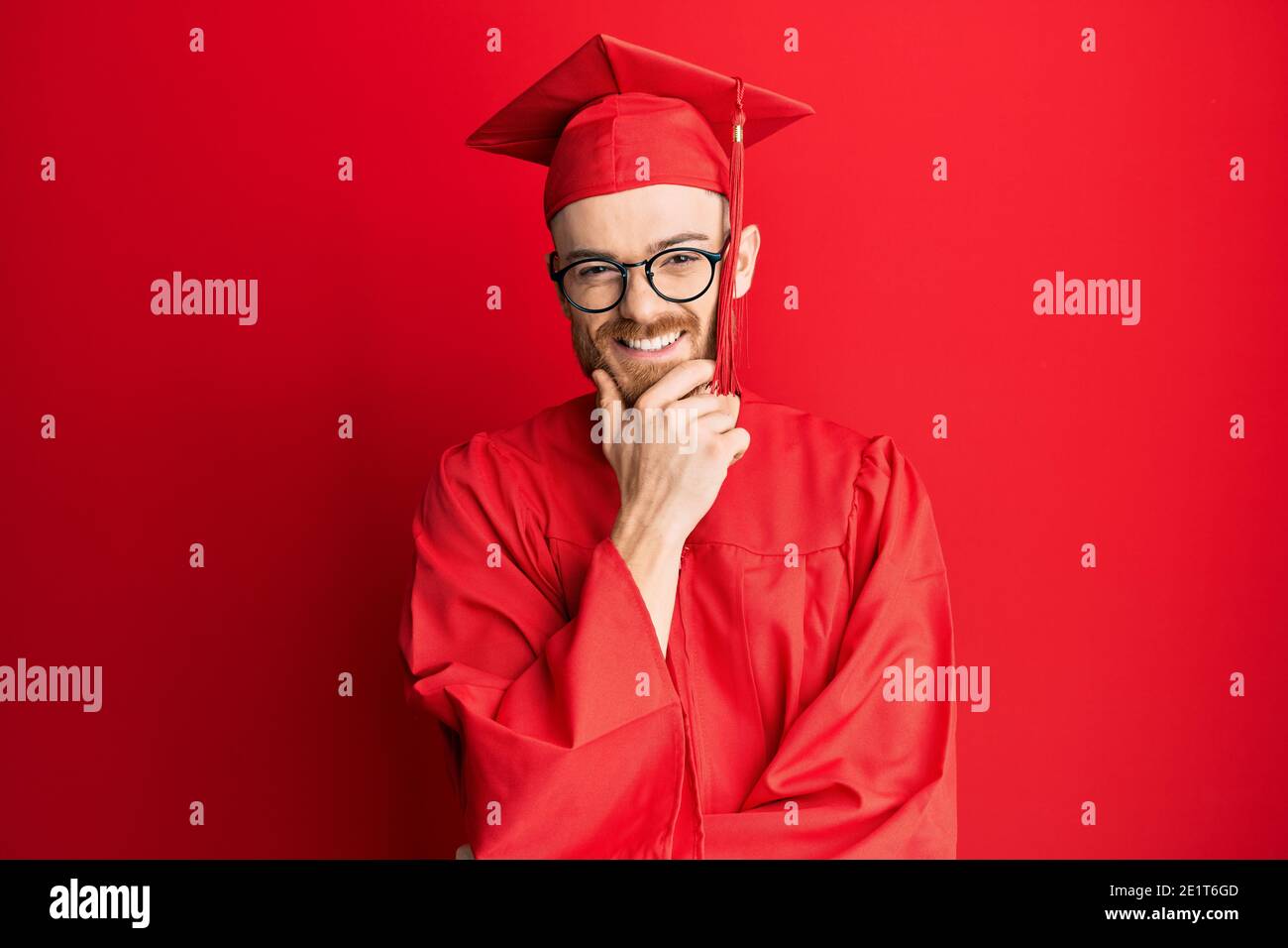 Young redhead man wearing red graduation cap and ceremony robe looking ...
