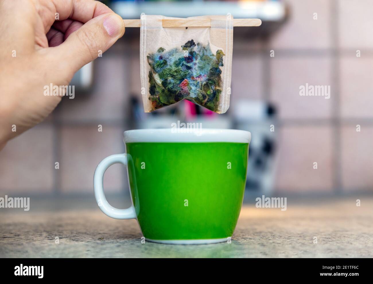 Hand holding a filter bag with wooden stick filled with herbal tea above a cup, kitchen background Stock Photo