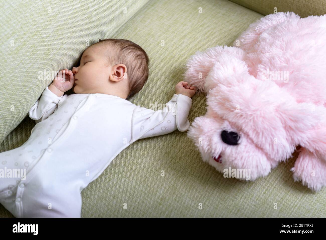 Baby sleeps in room, infant dreams on couch at home. Top view of sleeping small baby and pink soft toy on sofa. Little child in romper lying on his ba Stock Photo