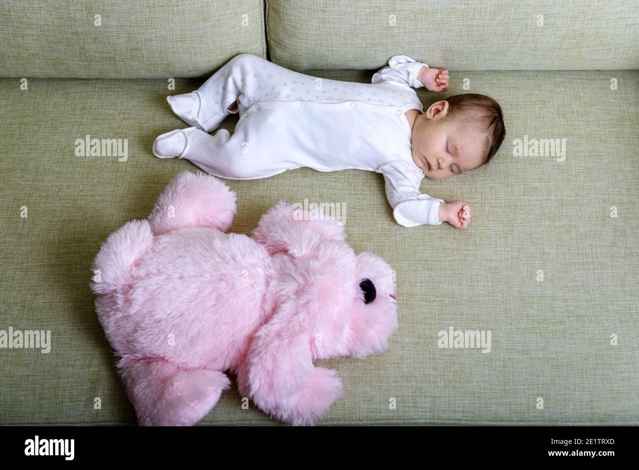 Baby sleeps in room, infant dreams on couch at home. Top view of sleeping small baby and pink soft toy on sofa. Little child in romper lying on his ba Stock Photo